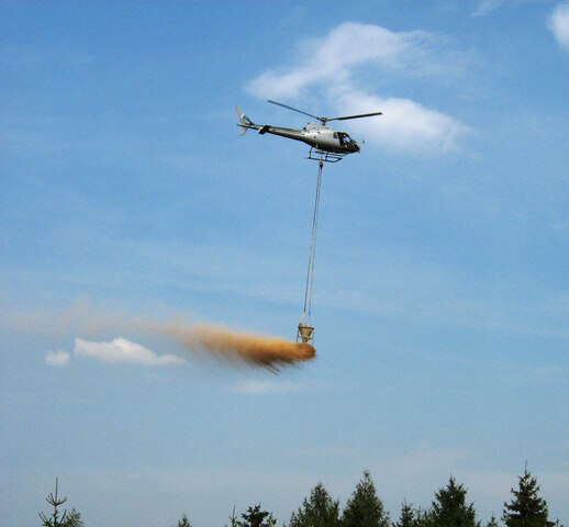 Helikopter fliegt mit geöffnetem Kalkbehälter über Baumkronen.