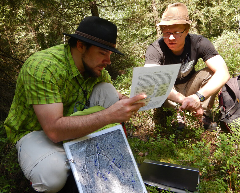 Ein Mitarbeiter hockt mit einem Besucher bei einer Führung im Gelände, um ihm etwas auf einem Blatt Papier zu zeigen.